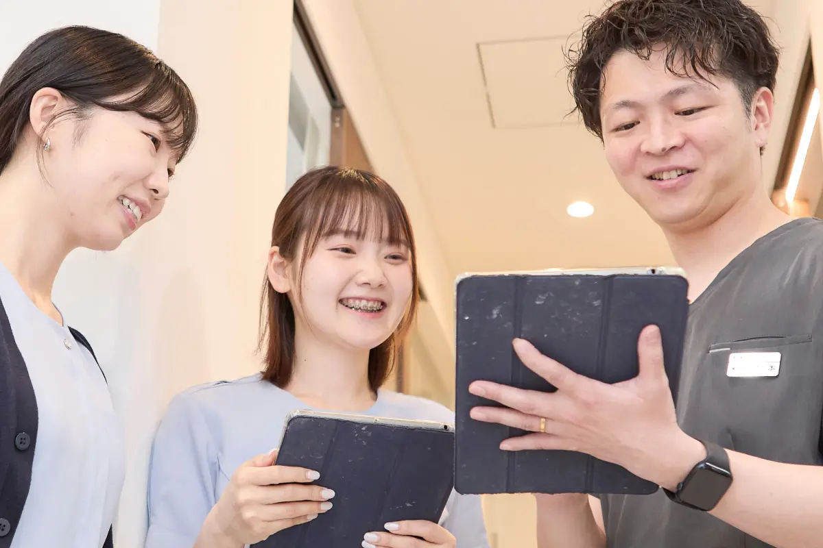 院長が笑顔でスタッフと談笑している、リラックスした雰囲気のポートレート。白衣を着ているが診療室ではなくスタッフルームや明るい廊下での自然な瞬間。「この先生と働きたい」と感じさせる温かみのある表情。