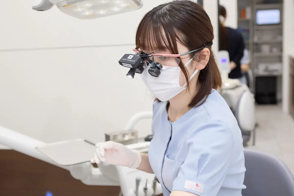 25歳前後の歯科衛生士がルーペを装着し、患者さんの口腔ケアに集中している横顔。スタイリッシュで技術的な雰囲気。診療室の清潔感と最新設備（口腔内スキャナー）が背景に見える。